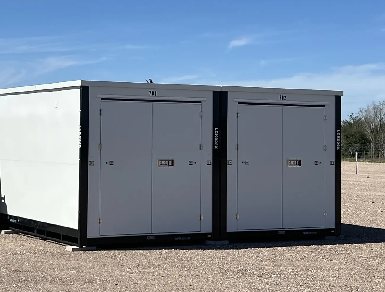 LockRig storage vaults 701 and 702: white steel units with double swing doors, sitting at ground level on the gravel yard