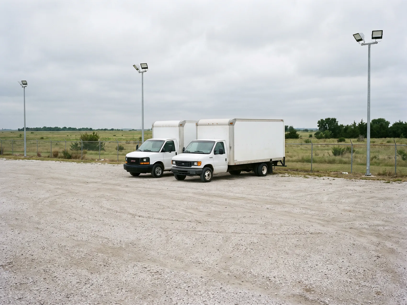 Two box trucks parked on a gravel lot beside a chain-link fence