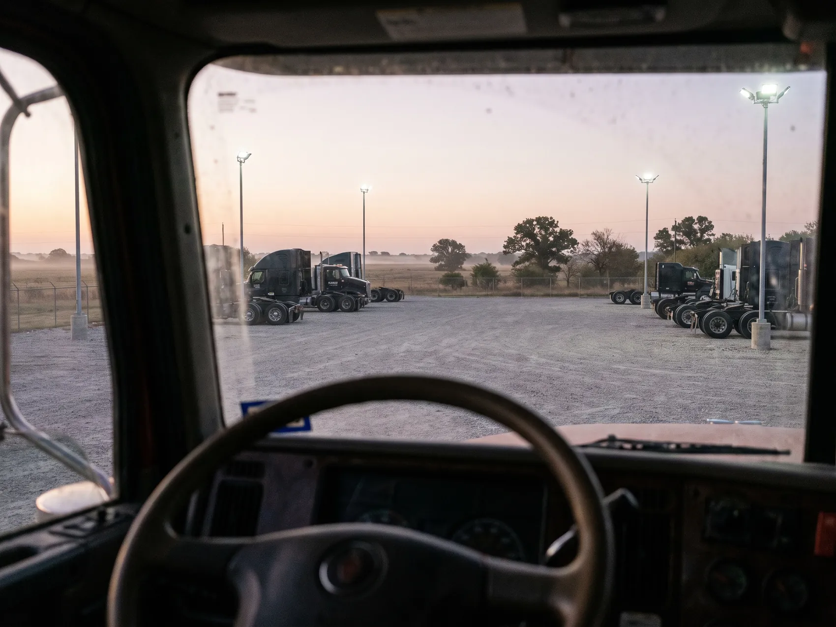 The view from a semi truck cab at dawn across a gravel lot of parked tractors