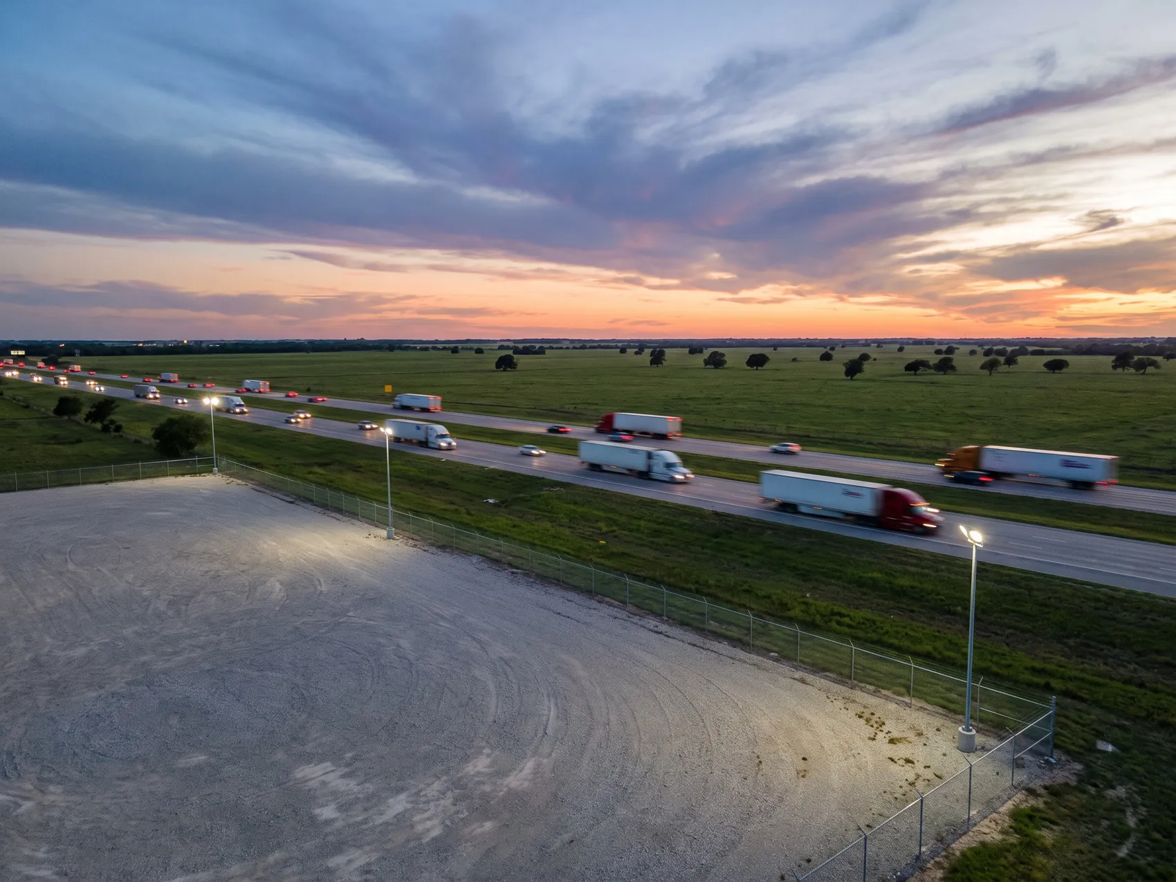 Semi trucks on a Texas interstate at dusk beside a lit gravel lot