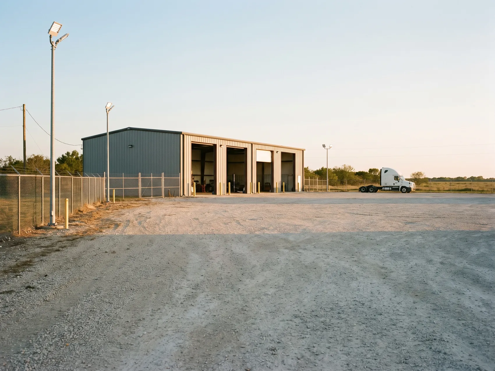 A gray steel building with roll-up bays beside a gravel yard at golden hour