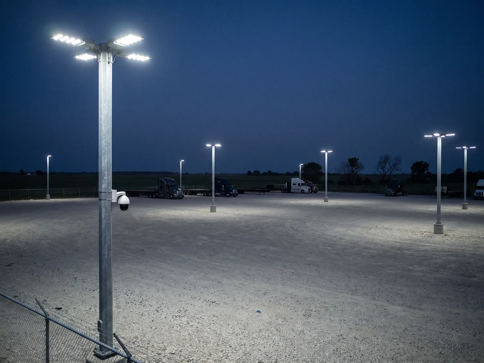 A gravel truck yard at night lit by LED floodlight poles, a security camera on the nearest pole
