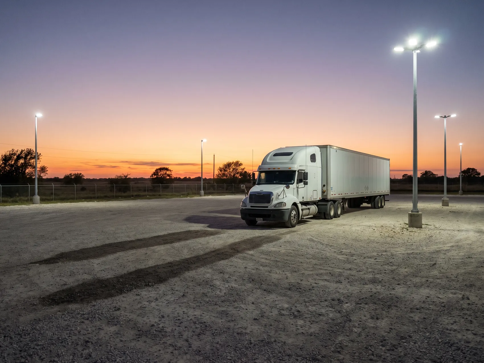 A semi tractor and 53-foot trailer parked on gravel at dusk under LED pole lights
