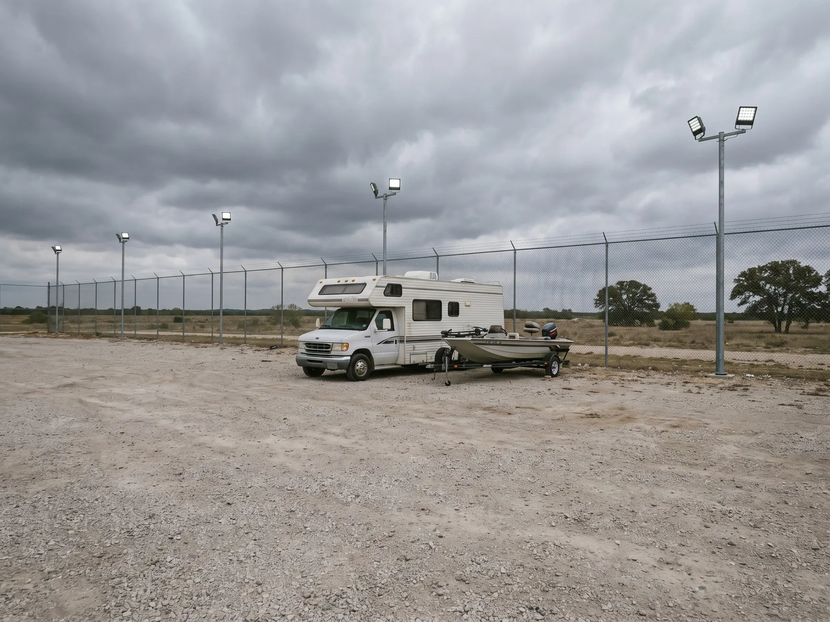 An RV and a boat on a trailer parked on dry gravel under a gray overcast sky